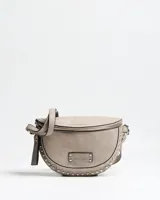 Riñonera de cuerina color gris taupe, con diseño semicircular, cierre superior y detalles de tachas metálicas en el borde inferior. Incluye correa ajustable y placa metálica con la inscripción de la marca.