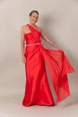 Vestido largo de fiesta color rojo, de corte recto y un hombro descubierto. Presenta una pieza de gasa que nace del hombro, formando el bretel y cayendo a lo largo del vestido.