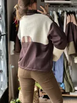 Buzo boxy marrón con diseño de rayas y guardas étnicas en tonos marrones, naranjas, blancos y negros en la parte superior y mangas, cuello alto y cierre frontal parcial con cremallera blanca, mangas largas con puños en tono crudo y corte holgado.