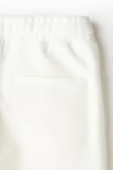 Pantalón tipo jogger de corte sastrero, color blanco, confeccionado en tejido de punto de peso medio. Presenta pliegue cosido frontal, cintura elástica con cordón de ajuste, bolsillos laterales y bolsillo trasero insertado.