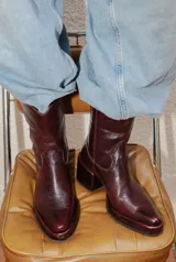 Bota estilo tejano de cuero color bordó con acabado brillante, taco cuadrado y punta cuadrada. Presenta costuras decorativas en el empeine y caña media.