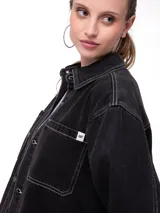 Camisa oversize de denim negro con costuras en contraste color blanco, bolsillos dobles en el pecho con solapa, cierre frontal de botones metálicos y dobladillo redondeado.