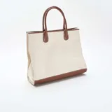 Bolso tipo tote de estructura rígida, confeccionado en lona color beige con detalles de cuero sintético en color marrón en la base, las asas y los bordes superiores.