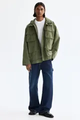 Campera estilo cargo de tejido ripstop, color verde militar, con cuello elevado que incluye capucha oculta. Presenta cierre frontal con solapa rompevientos, cuatro bolsillos delanteros con solapa y botón a presión, puños ajustables y cordón interno en la cintura.