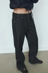 Jeans de tiro bajo y corte recto, confeccionados en denim negro con estampado de rayas verticales finas. Presentan cinco bolsillos, trabillas para cinturón y cierre frontal con cremallera y botón metálico.