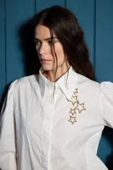 Camisa de corte clásico color blanco, con mangas largas abullonadas en los hombros y puños abotonados. Presenta un detalle de estrellas bordadas en color dorado sobre el lado izquierdo del pecho.