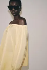 Blusa de lino color blanco, con escote recto ajustable mediante cordón y hombros descubiertos. Cuenta con manga larga abullonada terminada en elástico.