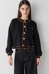 Cardigan de punto negro con detalles de costuras y bordes en color marrón. Presenta cuello redondo, cierre frontal con botones grandes y mangas largas con puños ajustados.