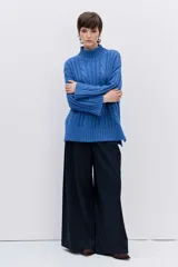 Sweater de punto con diseño de trenzas, cuello alto y mangas largas con puños acampanados. Presenta un corte holgado con terminación asimétrica en el ruedo.