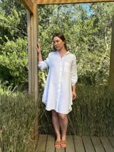 Vestido camisero blanco de lino, con cuello camisero, manga larga con botón para recoger, cierre frontal con botones de nácar, bolsillo en el pecho y diseño asimétrico más largo en la parte trasera.