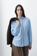 Camisa blanca de manga larga con detalles bordados en contraste a lo largo de la botonadura, el cuello y los puños. Presenta una silueta relajada y un diseño clásico con un toque artesanal.