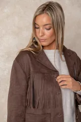Chaqueta de gamuza sintética color marrón, con diseño de flecos en el pecho y la espalda. Presenta cierre frontal con botones, cuello camisero y bolsillos cuadrados frontales.