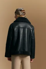 Campera tipo gamulán de cuero sintético negro, con interior y detalles de corderito sintético al tono en cuello y ruedo. Presenta cierre frontal metálico y diseño de silueta relajada.