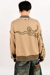 Sweater tejido reversible de corte boxy fit, con cuello redondo y puños acanalados. Presenta un diseño gráfico abstracto de estilo gótico que recorre el pecho y las mangas. Un lado es de color marrón oscuro con detalles en beige, y el otro lado es de color beige con detalles en marrón oscuro.