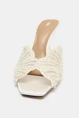 Sandalia de tacón alto estilo mule, de color blanco crudo, con diseño en la pala en forma de concha decorada con perlas y cuentas brillantes, punta cuadrada y tacón fino.