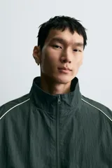 Chaqueta técnica de corte relajado en color verde oscuro, con cuello subido, cierre frontal de cremallera y detalles de líneas en contraste. Presenta bolsillos frontales con cremallera oculta y acabados elásticos en puños y ruedo.