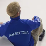 Campera deportiva Adidas de la selección argentina, color azul con cierre frontal, cuello alto, puños y cintura acanalados. Presenta las icónicas tres tiras blancas en las mangas, el logo del trébol de Adidas en el pecho derecho y el escudo de la AFA en el pecho izquierdo.