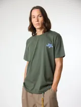 Camiseta de manga corta color verde militar, de corte regular fit y confeccionada en algodón orgánico. Presenta un estampado gráfico grande en la espalda con el texto 'blue banana' en tonos celestes y oscuros, acompañado de un pequeño recuadro blanco con inscripciones.
