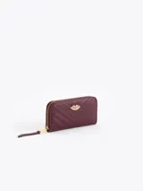 Maxi billetera de cuero vacuno con diseño matelassé en forma de V. Presenta un aplique metálico dorado en forma de labios en el frente y cierre perimetral con tirador de cuero.