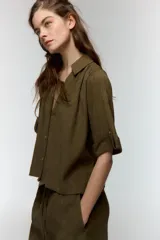 Camisa de cuello solapa y escote en pico, con mangas cortas que incluyen trabilla y botón. Presenta un cierre frontal con botones y un diseño de corte recto.
