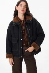 Chaqueta de largo medio y silueta holgada, confeccionada en tela de jean color negro con costuras a contratono. Presenta forro interno de corderito color marrón, cuello alto, cierre frontal con cremallera y botones a presión, y bolsillos plaqué con tapa en el frente.