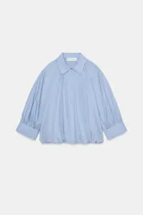 Camisa de algodón con cuello solapa, mangas abullonadas por debajo del codo y bajo acabado en globo. Presenta cierre frontal con botones ocultos.