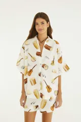 Camisa oversize blanca con estampado de instrumentos musicales, cuello camisero, botones frontales, hombros caídos y mangas cortas amplias.