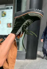 Borcegos de caña alta color verde oscuro, confeccionados en gamuza y terciopelo, con cordones de terciopelo y suela de goma negra con detalle naranja.