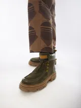 Borcegos de caña baja con diseño robusto, confeccionados en una combinación de gamuza y cuero sintético en tonos verde oliva. Presentan suela track de goma dentada en color suela, cordones al tono con ojalillos amarillos y cuello acolchado para mayor confort.