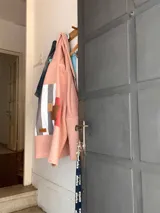 Campera de corderito sintético color rosa, con capucha y diseño de collage en la espalda que combina franjas verticales en tonos grises, blanco y parches en texturas de marrón y terracota. Presenta un corte holgado y mangas anchas.