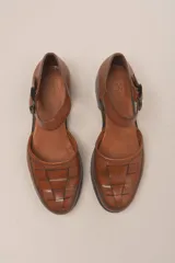 Zapato de cuero color borgoña, con puntera redondeada y diseño trenzado. Presenta una tira sobre el empeine con hebilla metálica negra.