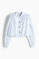 Camisa corta de sarga de algodón en color celeste, con cuello elevado, botonadura frontal con botones oscuros y corte trapecio. Presenta hombreras, mangas largas abullonadas con puños abotonados y detalles de frunces en los hombros.