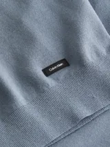 Sweater de punto liso color azul grisáceo, con cuello redondo y detalles de canalé en cuello, puños y bajo. Presenta una pequeña etiqueta con el logo de Calvin Klein sobre el bajo.