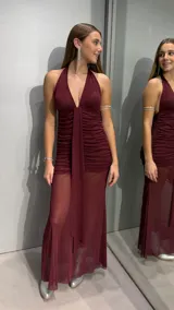 Vestido largo color bordó con escote halter profundo y diseño fruncido en el torso. La falda presenta una capa superior opaca con un panel central drapeado y una capa inferior transparente que aporta movimiento y elegancia.