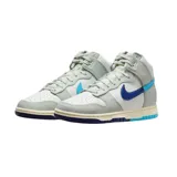Championes Nike Dunk High Retro SE de corte alto, con diseño en paneles de cuero blanco y gris. Presenta un swoosh lateral dividido en dos tonos de azul, suela intermedia con acabado envejecido y suela exterior bicolor en azul claro y oscuro.