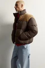 Cazadora puffer relaxed fit color marrón con canesú superior en tono tostado de acabado aterciopelado. Cuenta con cuello subido, manga larga, bolsillos de vivo en la cadera, acabados en elástico y cierre frontal con cremallera.