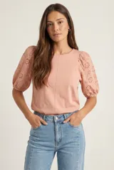 Remera de algodón color rosado con mangas cortas abullonadas de broderie con diseño floral calado. Presenta cuello redondo y costura vertical central en el frente.