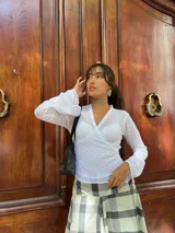 Remera blanca de encaje con diseño cruzado tipo wrap, mangas largas con terminación acampanada y detalle de bordado floral a tono. Presenta un ajuste al cuerpo con lazo lateral y borde con terminación delicada en el escote.