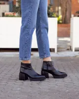 Botín de cuero negro, estilo botineta, con taco ancho y plataforma. Presenta un diseño liso y brillante, con cierre lateral.