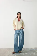 Sweater de tejido bouclé color crudo con detalles de ribete en color rojo en el cuello, puños y bolsillos. Presenta un diseño de manga larga, cuello en V y bolsillos frontales.