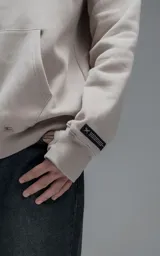 Hoodie oversize de color beige, confeccionado en frisa premium. Presenta capucha ajustable con cordones, bolsillo tipo canguro frontal y puños con aberturas para pulgares.