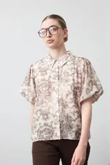 Camisa de manga corta abullonada, con cuello camisero y botones frontales. Presenta un estampado de inspiración barroca en tonos marrón y beige claro.