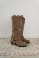 Bota estilo tejana de caña alta, confeccionada en cuero marrón con acabado texturizado. Presenta punta cuadrada, taco bajo y una correa con hebilla metálica ajustable y estribo desmontable en la zona del tobillo.