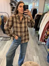 Camisa de manga larga con estampado de cuadros escoceses en tonos marrones y beige, confeccionada en tela suave con caída. Presenta cuello clásico, cierre frontal con botones y detalles de flecos de cuero rojo aplicados.