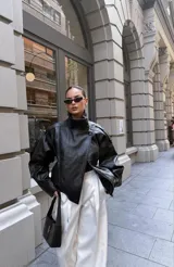 Chaqueta negra de efecto cuero, estilo bomber o motociclista, con cuello alto abotonado y cierre frontal asimétrico. Presenta detalles de costuras verticales y hombreras pequeñas. El bajo es elástico.