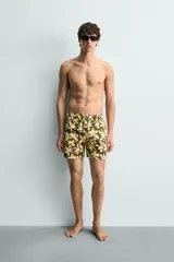 Bañador de hombre con estampado floral en tonos amarillo y blanco sobre fondo verde oliva. Presenta cintura elástica ajustable con cordón, bolsillos frontales y forro interior.