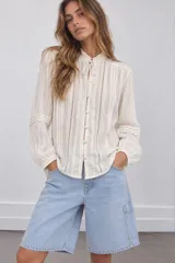 Blusa de tejido texturizado color blanco, con cuello alto con volados y cordón para atar, cierre frontal de botones y mangas largas abullonadas con detalles de encaje y tablas verticales.