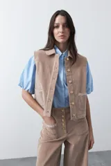 Chaleco de pana color marrón oscuro, con cuello camisero, cierre frontal de botones metálicos dorados y dos bolsillos frontales con detalle de bordado geométrico.