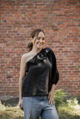 Blusa asimétrica de lino color negro, con un hombro al descubierto y detalle de volado en el escote. Presenta manga larga abullonada y corte recto.