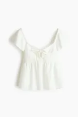 Blusa corta blanca en punto pointelle con textura, escote corazón, mangas cortas circulares, detalle cut-out con cordón de ajuste en el busto y diseño acampanado con costura fruncida.